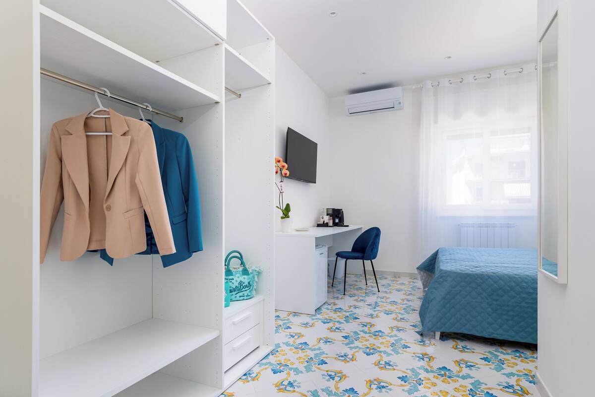 Une chambre à coucher avec un lit, un bureau et une chaise. On y trouve une armoire dans laquelle sont suspendus des vêtements.