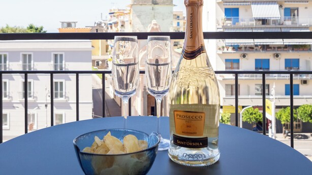 Una mesa con una botella de Prosecco y dos copas en un balcón con vista a un paisaje urbano.