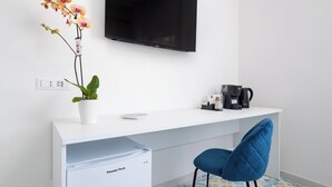 Habitación de hotel moderna con un escritorio blanco, un televisor de pantalla plana, cafetera, un refrigerador pequeño, una silla azul y una orquídea en maceta.