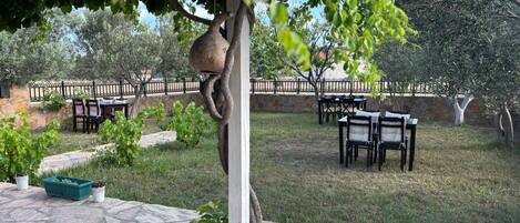Un área de estar al aire libre con suelo de piedra, mesas y sillas, rodeada de vegetación y un muro de piedra.