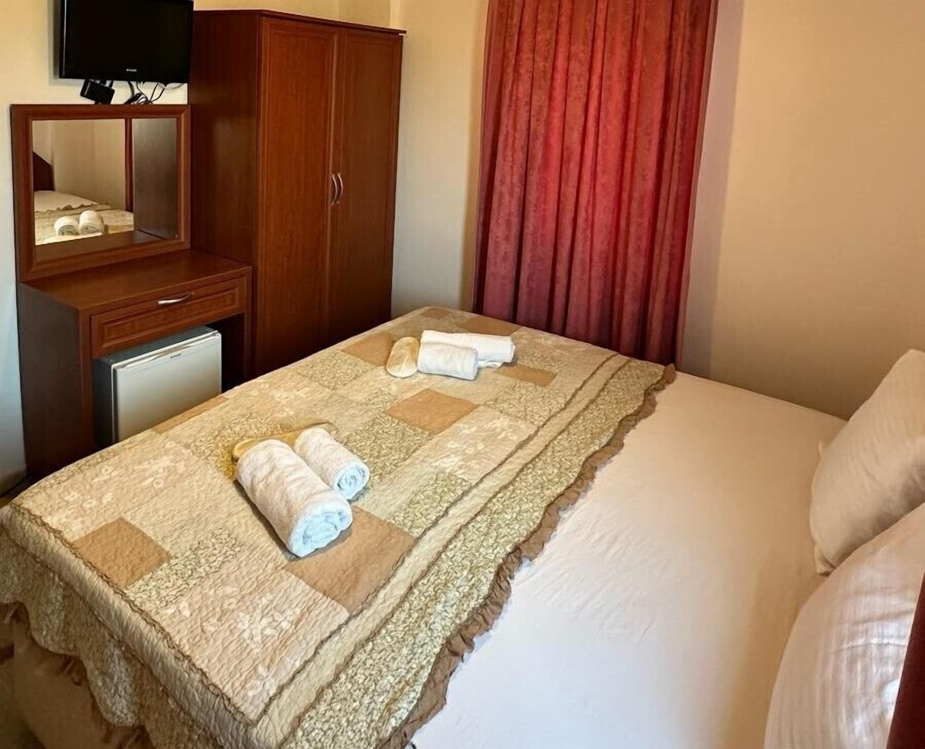 A hotel room with a bed, a wooden wardrobe, a small refrigerator, and a television mounted on the wall.