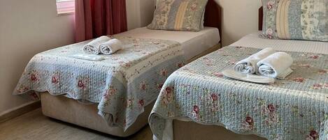 Duas camas de solteiro com roupa de cama e toalhas florais, uma janela com cortinas vermelhas e um chão de madeira claro.
