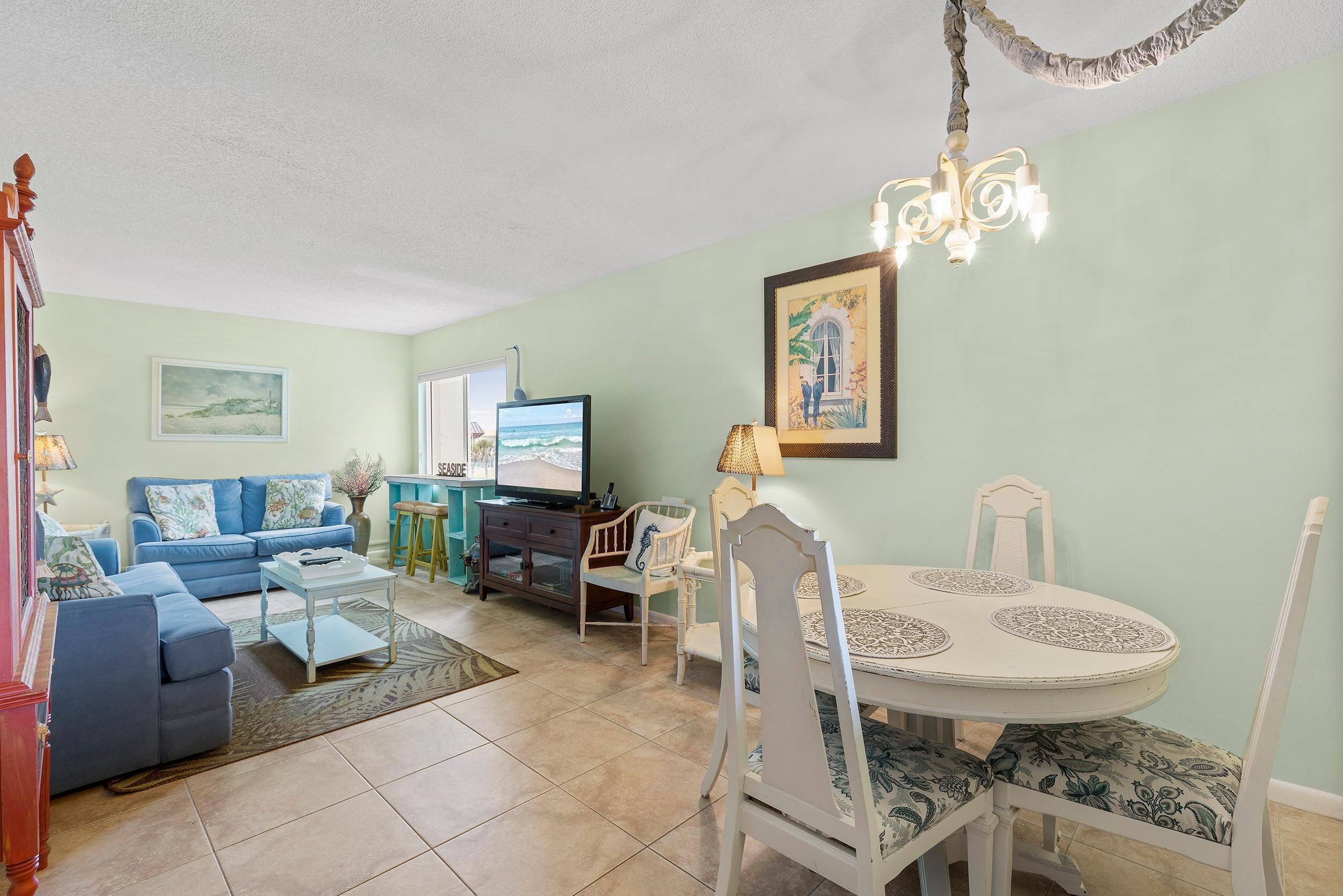 A living room with a blue sofa, a dining table with white chairs, a TV, and a chandelier.