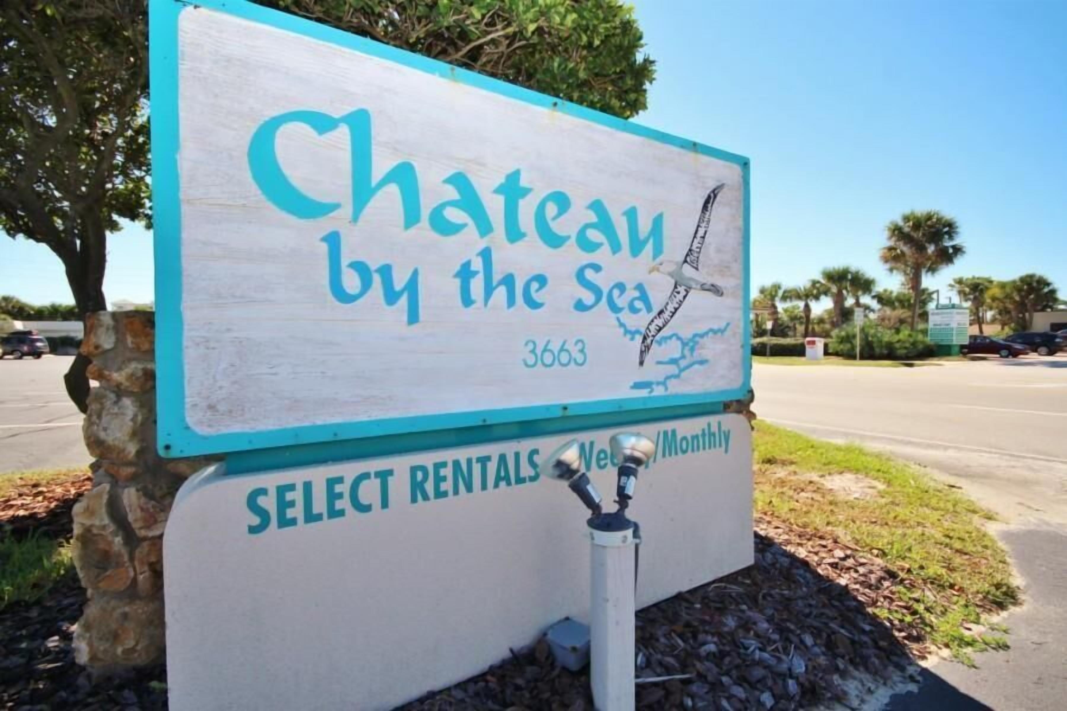 A sign for 'Chateau by the Sea' with a beach view illustration, indicating a beachfront property. The sign also mentions 'Select Rentals' and 'Weekly/Monthly' options.