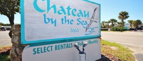 A sign for 'Chateau by the Sea' with a beach view illustration, indicating a beachfront property. The sign also mentions 'Select Rentals' and 'Weekly/Monthly' options.