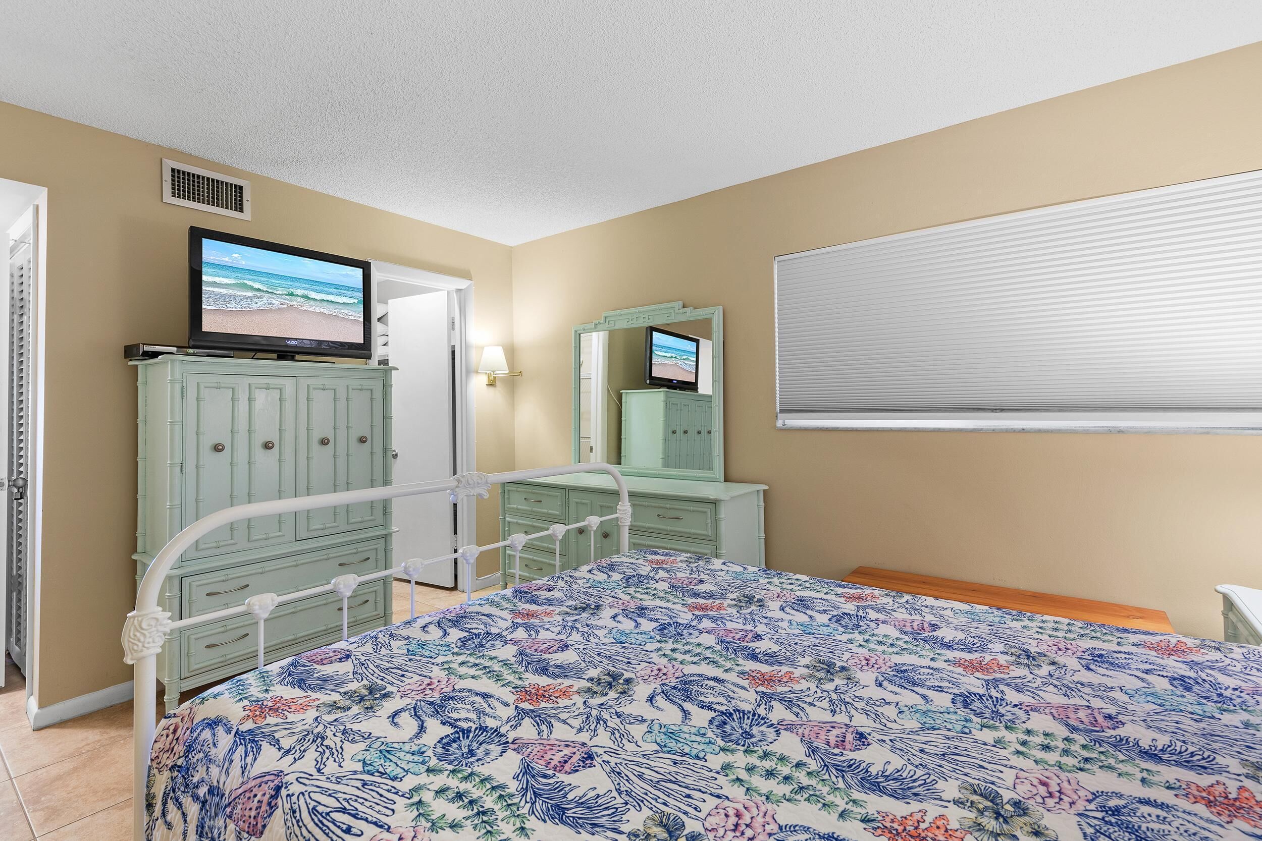 A bedroom with a bed, a television on a dresser, a mirror, and a window with blinds.