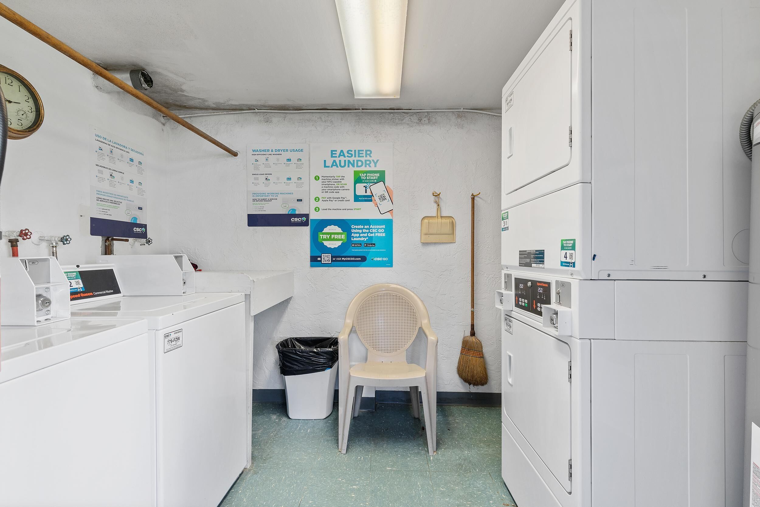 A laundry room with a washing machine, dryer, and a chair. There are instructional posters on the wall and a broom hanging on the wall.