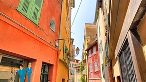 A narrow street with colorful buildings, including a red building with shutters and a yellow building with a balcony. - N&J - REFLET D'OR - Vieux Nice - Neau beach (Niza)