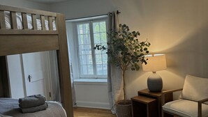 Een slaapkamer met een stapelbed, een nachtkastje met een lamp, een stoel, een kamerplant en een raam met gordijnen.