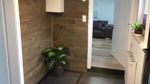 Un couloir avec un mur en bois, une plante en pot et, au loin, on aperçoit un salon avec un canapé et une plante.