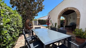 An outdoor patio with a glass-top table and chairs, surrounded by greenery and a white building with an archway. - El Rincón del Montgó - private swimming pool (Xàbia)