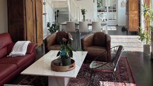 A spacious living room with a red sofa, a coffee table, and a patterned rug. There is a staircase leading to an upper level, and a dining area in the background.