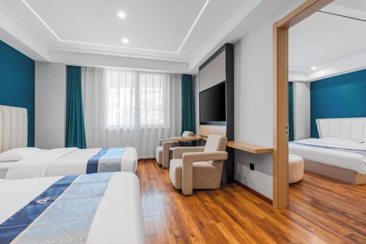A modern hotel room with a wooden floor, a large bed, a sofa, a TV mounted on the wall, and a seating area with chairs.