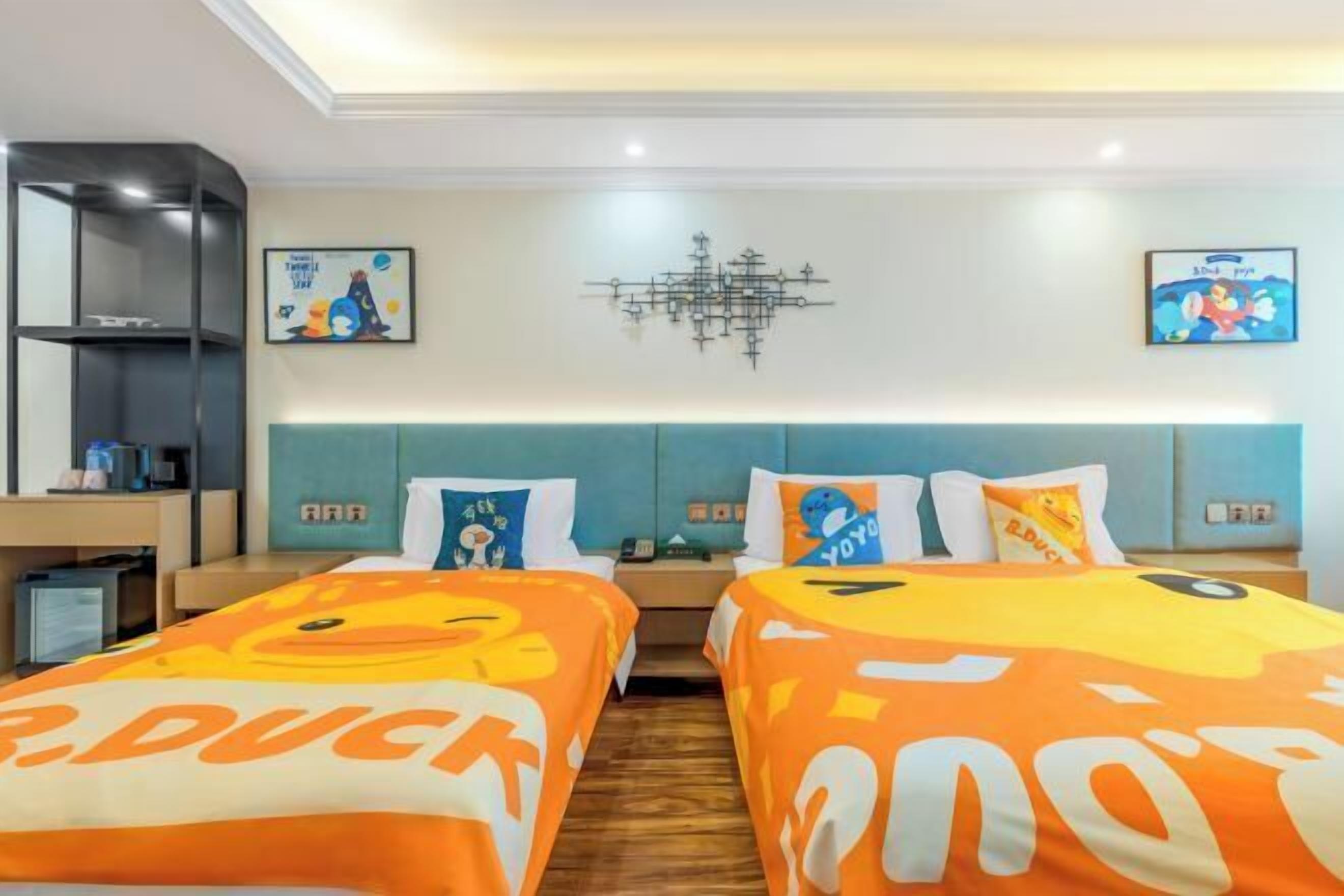 A hotel room with two beds, a desk, a bookshelf, and artwork on the walls.