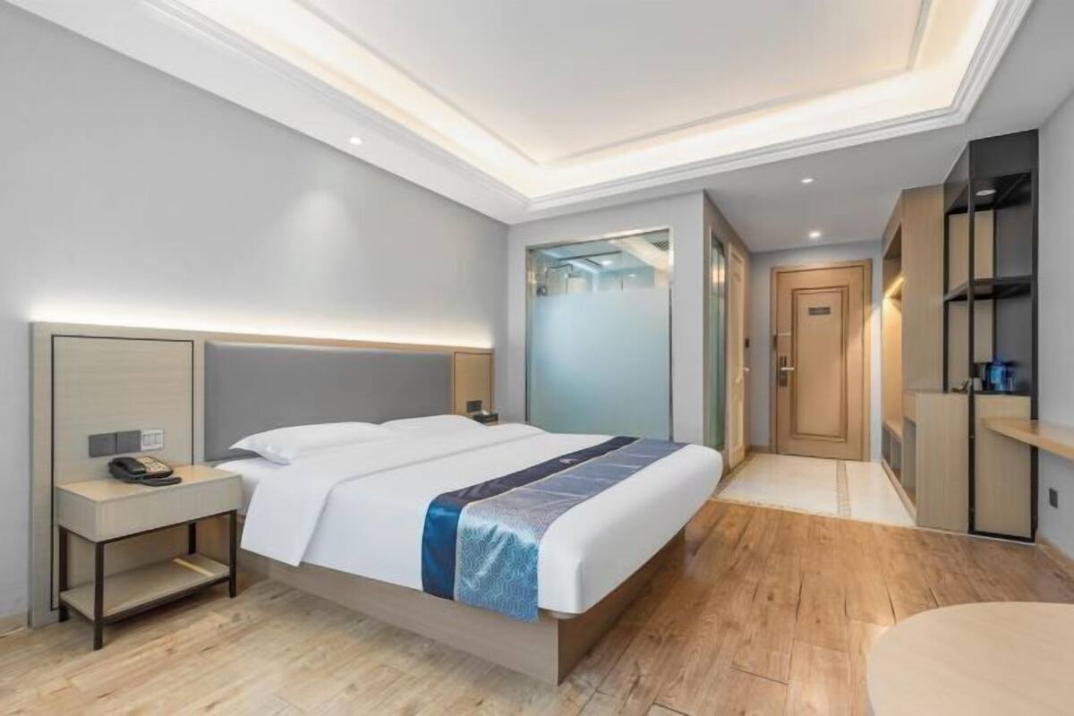 A modern hotel room with a large bed, bedside tables, a wardrobe, and a bathroom visible through a glass door.