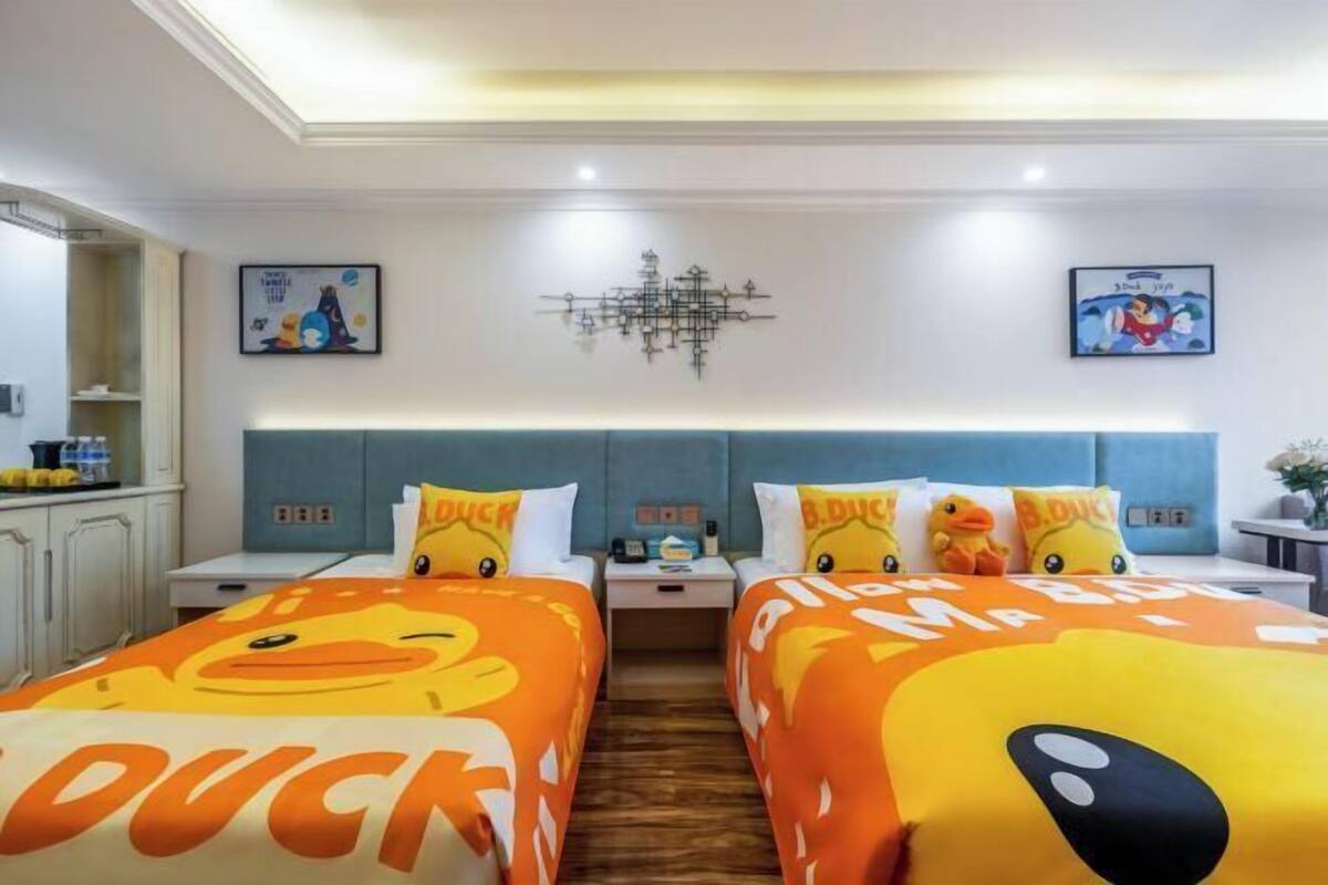 A hotel room with two beds, a chandelier, artwork on the walls, and a kitchenette.