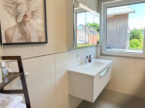 A modern bathroom with a white vanity, a large mirror, and a window offering a view of buildings and trees.
