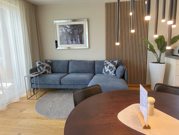 A modern living room with a grey sofa, a round wooden table, a framed black and white photo on the wall, and a vertical wooden panel.