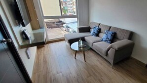 Una sala de estar moderna con un sofá color beige, dos cojines estampados, una mesa de centro de vidrio y vistas a una obra en construcción a través de un ventanal amplio.