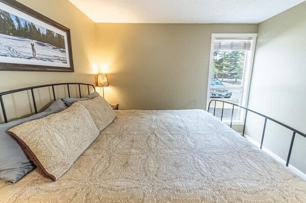 A bedroom with a bed, a nightstand, a lamp, a window with a view of a car and trees, and a framed picture on the wall. - Walking distance to shops, brewery and eateries. (Sunriver)