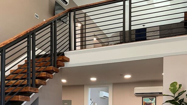 Un vaste espace de vie comprenant un canapé gris, une table basse en bois, un escalier avec une rampe noire et une plante en pot.