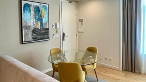Ένα μοντέρνο σαλόνι με ένα τραπέζι από γυαλί και κίτρινες καρέκλες, έναν μπεζ καναπέ, έναν πίνακα στον τοίχο και μια πόρτα με διακόπτη φωτισμού.