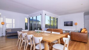 Un vaste espace de vie comprenant une table à manger en bois, des chaises blanches, un canapé en cuir marron et un téléviseur fixé au mur.