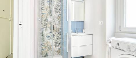 A bathroom with a glass shower enclosure, a white sink, a mirror, and a front-loading washing machine.