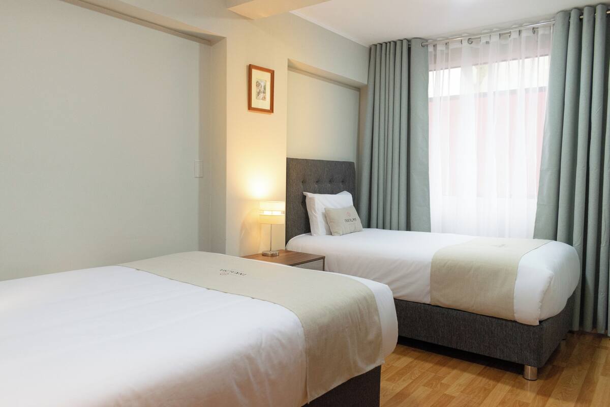 A hotel room with two beds, a bedside table with a lamp, a framed picture on the wall, and a window with curtains.
