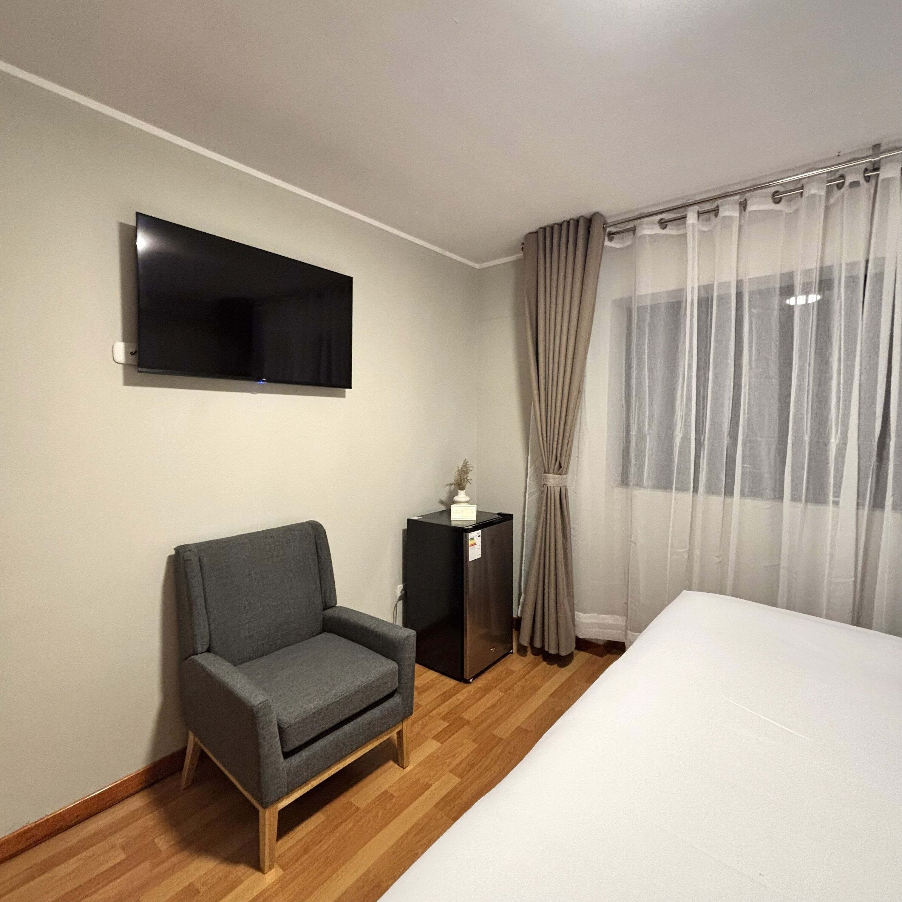 A hotel room with a bed, a grey armchair, a TV mounted on the wall, a small table with a lamp, and sheer curtains on the window.