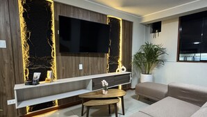A modern living room with a large flat-screen TV, a wooden accent wall, a white console table, a round coffee table, and a beige sofa. - HOTEL INKALMA SANTA MARTA (Arequipa)