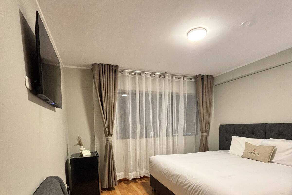 A hotel room with a bed, a TV mounted on the wall, a small table with a lamp, and a chair.