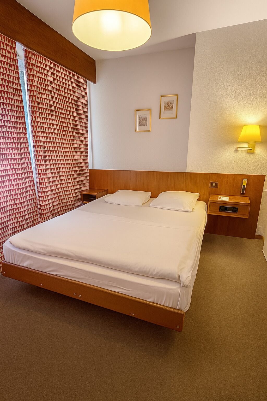 Une chambre d’hôtel avec un grand lit, une tête de lit en bois, une table de chevet, une lampe et un rideau à motifs.