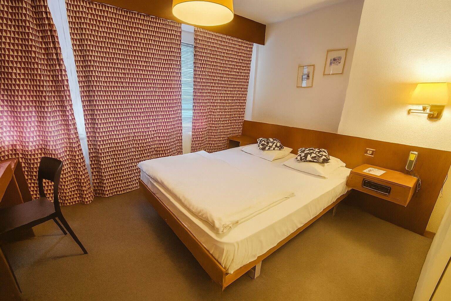 A hotel room with a bed, a desk, a chair, a nightstand, and a lamp.