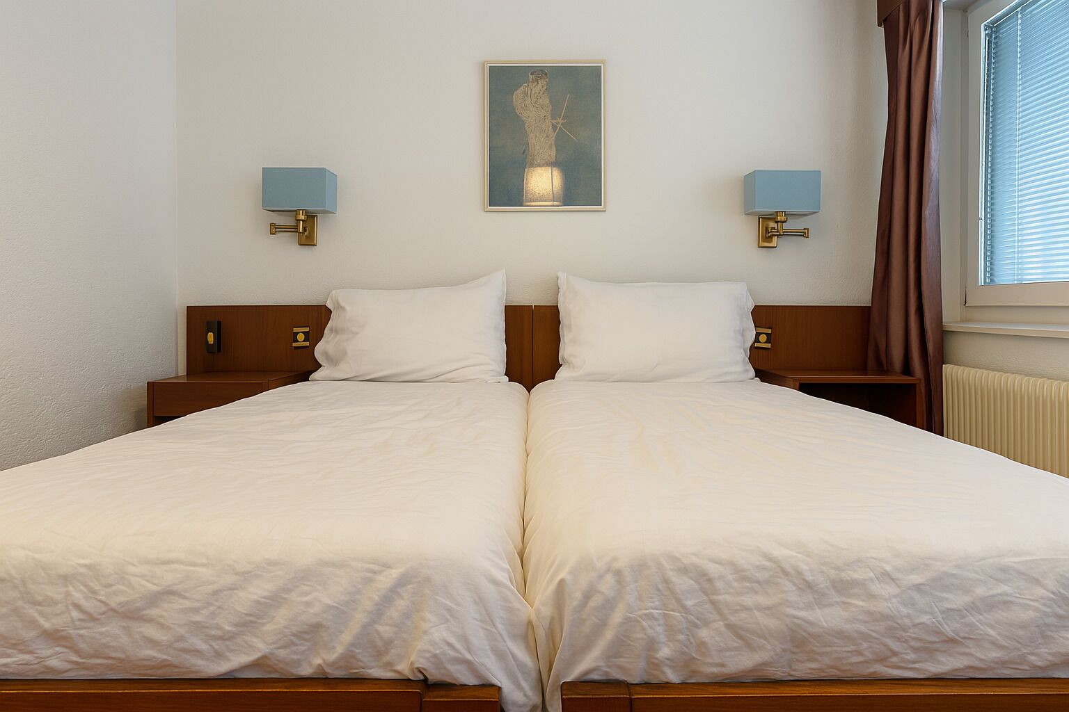 A double bed with white linens, two bedside tables with lamps, a framed artwork above the bed, and a window with curtains.