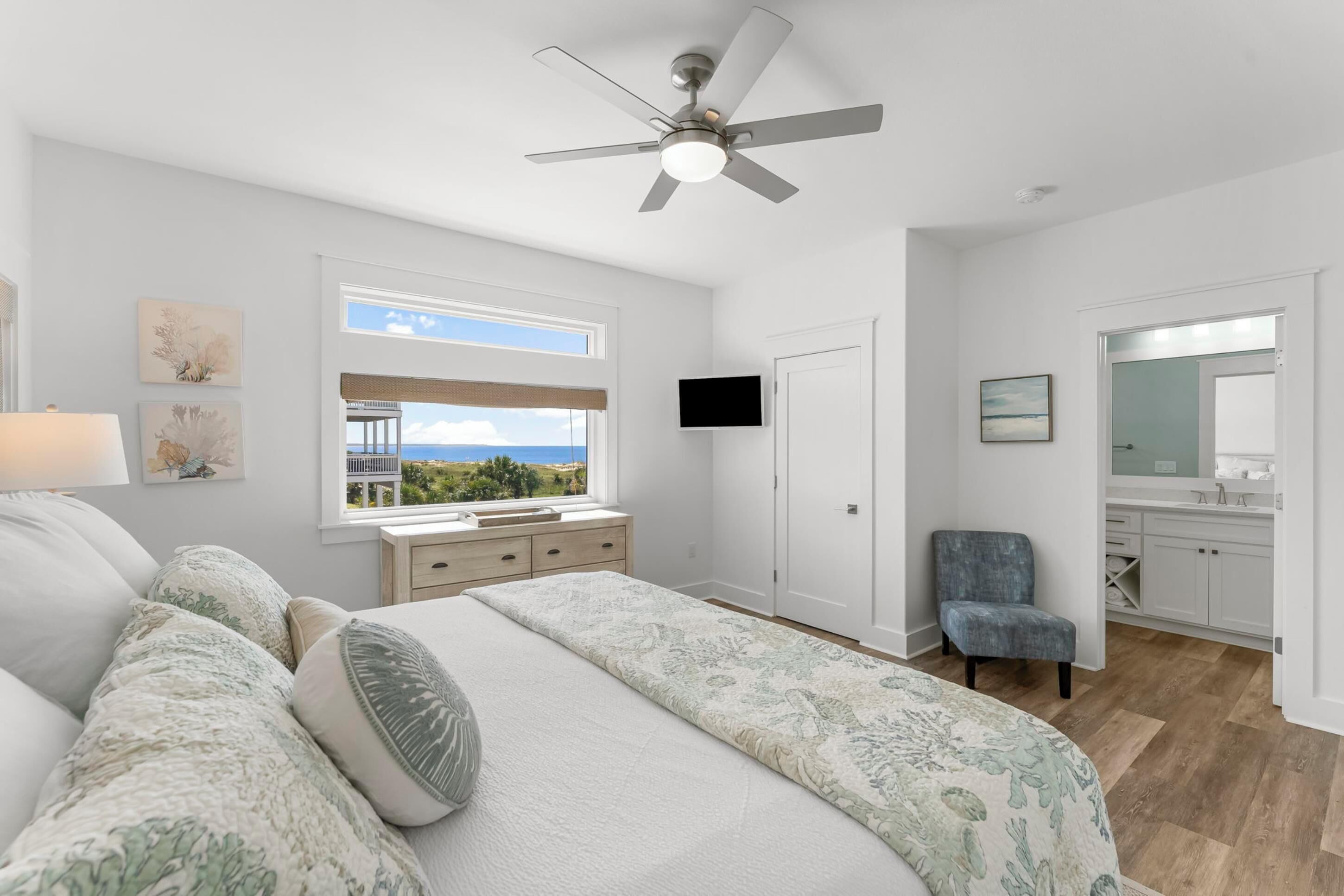 Un dormitorio con cama, ventilador de techo, ventana con vistas, televisor, una silla y baño con espejo y lavabo.