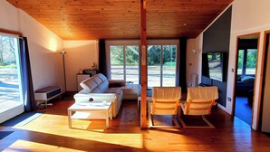 A spacious living area with a wooden ceiling, large windows, a sofa, chairs, and a coffee table. - Guesthouse in wooded grounds (Saint-Vincent-de-Tyrosse)