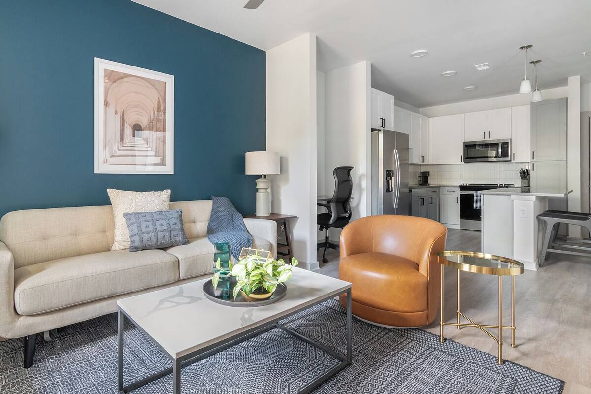 A modern living room with a sofa, coffee table, and armchair, a dining area with a table and chairs, and a kitchen with appliances.