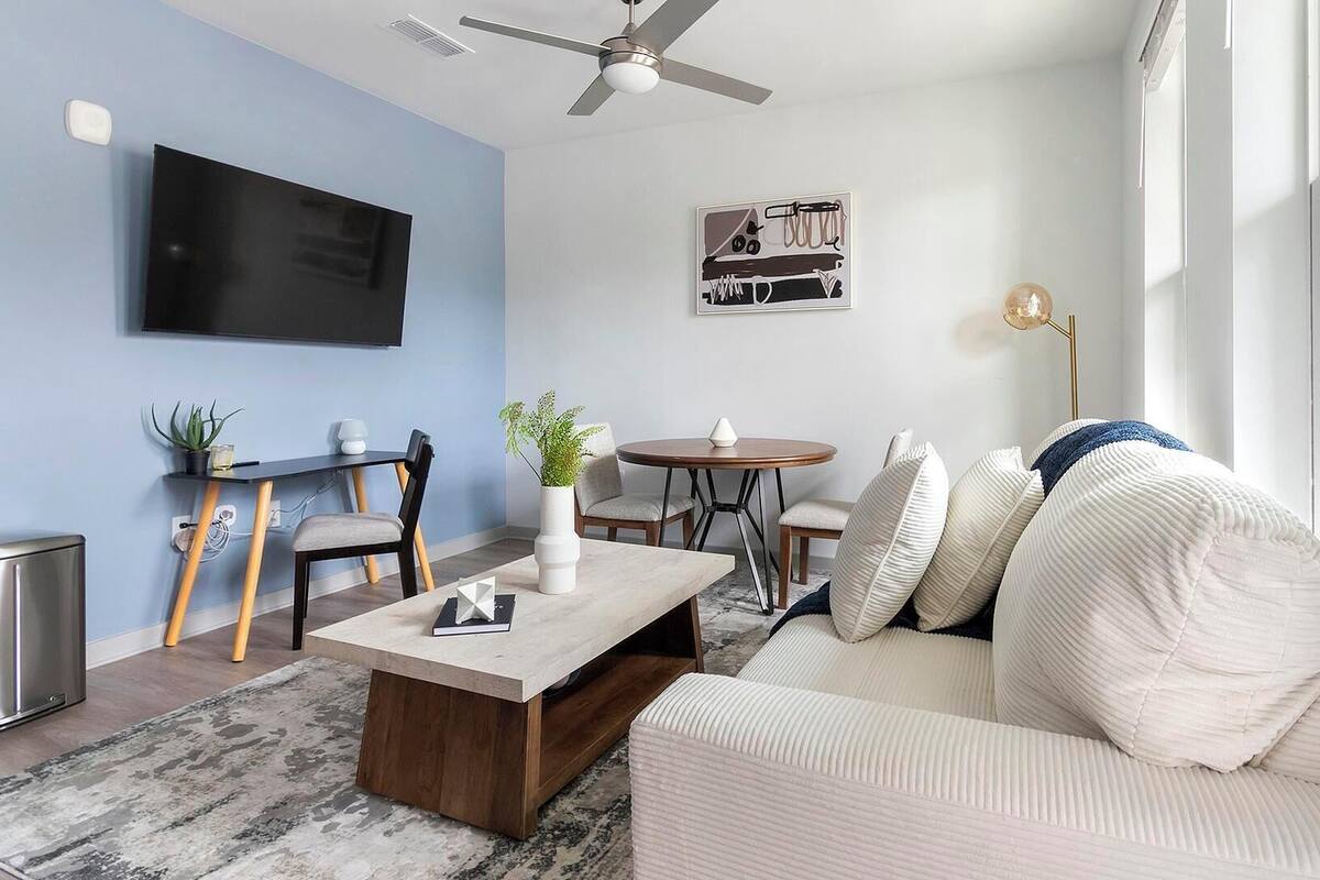 A modern living room with a flat-screen TV, a ceiling fan, a dining area with a round table, and a sofa with decorative pillows.