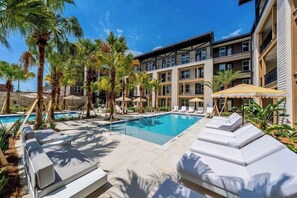 A pool area with lounge chairs, palm trees, and a building with multiple balconies. - Historic Springfield Apartments by Landing (Jacksonville)