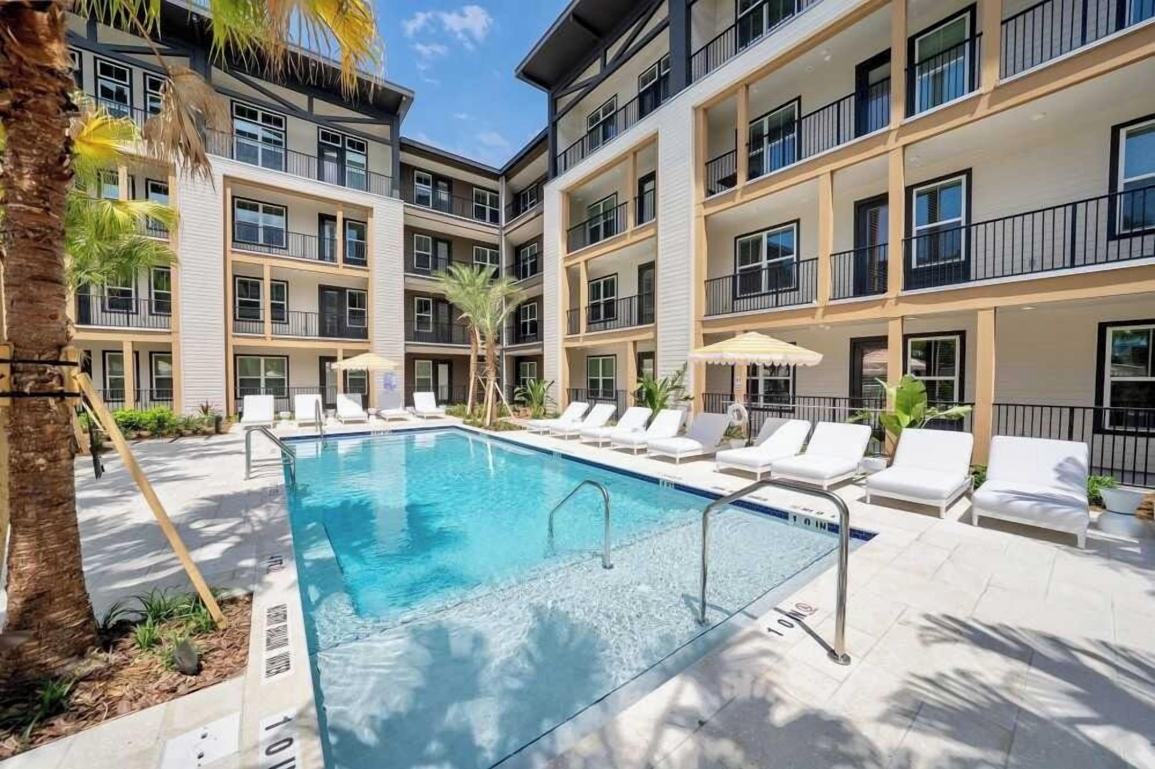 A pool area with lounge chairs and a clear blue pool, surrounded by multi-story buildings with balconies.
