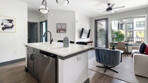 A modern kitchen with a dishwasher, a dining area with a blue chair, and a living room with a sofa and a plant. - Southbank Apartments by Landing (Jacksonville)