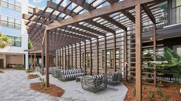 A covered outdoor seating area with wooden beams and metal furniture, surrounded by a brick building and plants. - Southbank Apartments by Landing (Jacksonville)