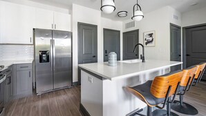 A modern kitchen with a stainless steel refrigerator, a built-in coffee machine, a large island with a sink, and bar stools. - Southbank Apartments by Landing (Jacksonville)