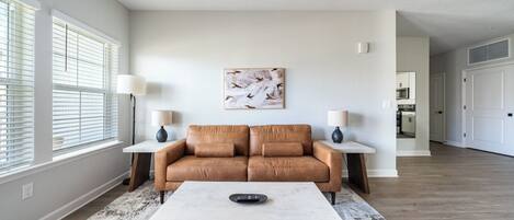 A spacious living room with a brown leather sofa, a white coffee table, and two side tables with lamps. A large abstract painting hangs on the wall, and there is a view into the kitchen through an open door.