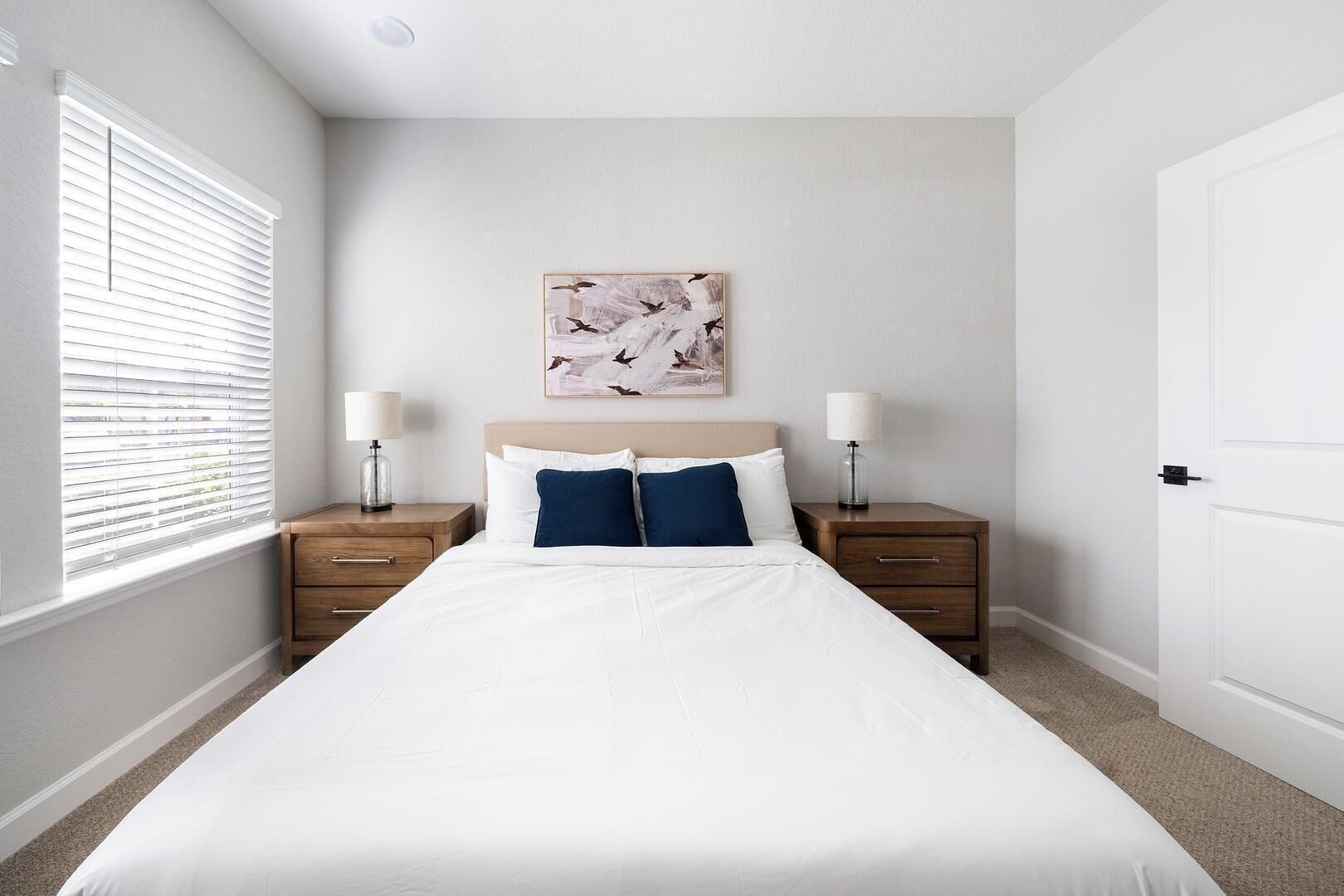 A bedroom with a large bed, two bedside tables, a nightstand, a window with blinds, and a wall-mounted artwork.