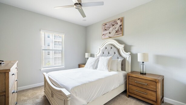 Une chambre à coucher comprenant un lit, des tables de chevet, un ventilateur de plafond, une fenêtre avec des stores et un tableau accroché au mur.