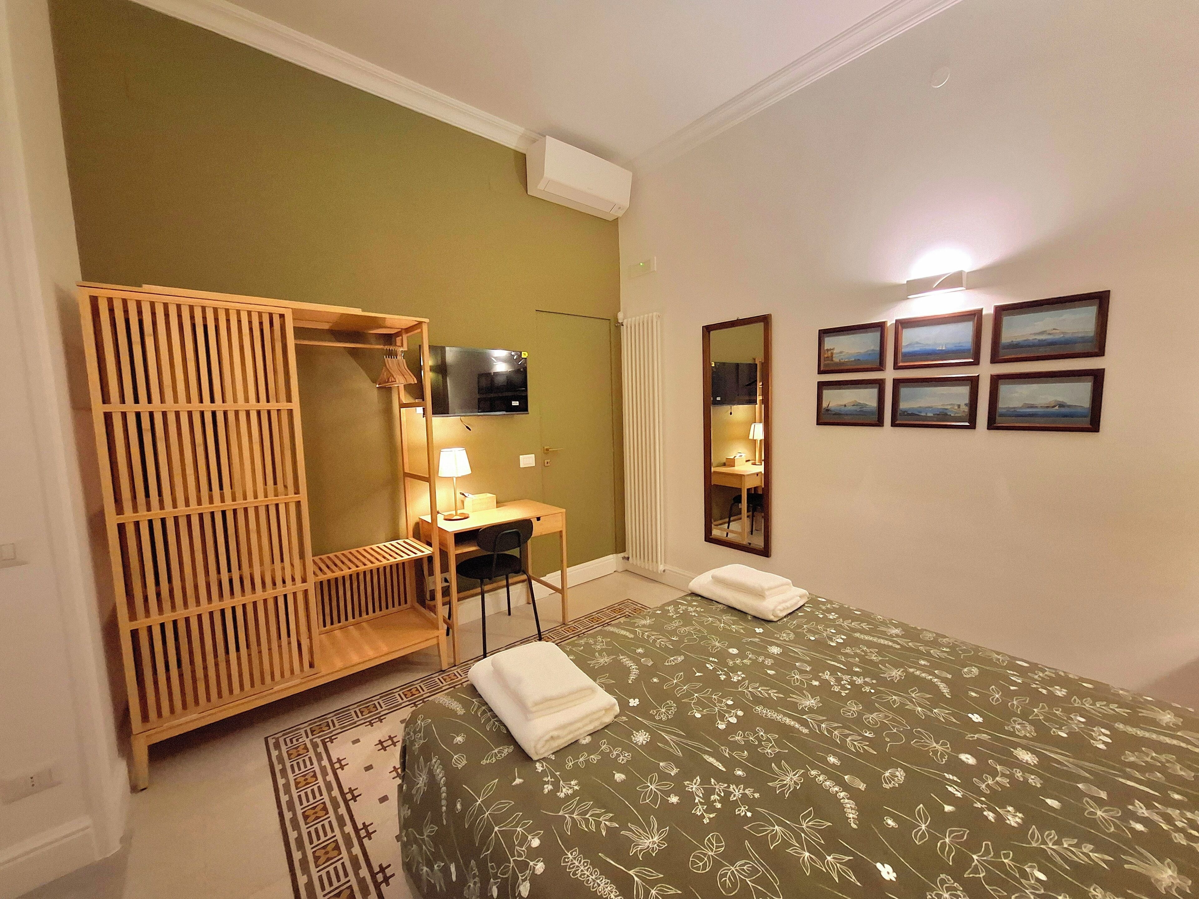 A hotel room with a bed, a desk, a TV, a mirror, and framed pictures on the wall.