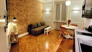 A modern kitchen with a built-in oven, a dining area with a white table and chairs, a sofa with a patterned wall behind it, and a wooden herringbone floor. - TALISMANO LUXURY MAISON (Napoli)