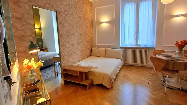 A hotel room with a bed, a small table with a vase of flowers, a chair, a window with curtains, and a radiator. - TALISMANO LUXURY MAISON (Napoli)